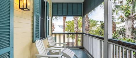A porch with white Adirondack chairs, a ceiling fan, and a view of palm trees and a house.