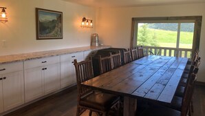 A dining area with a long wooden table, white cabinets, and a view of a green landscape through a window. - Charming farmhouse property on 20 acres of land, comfortably sleeps 13   (Lava Hot Springs)
