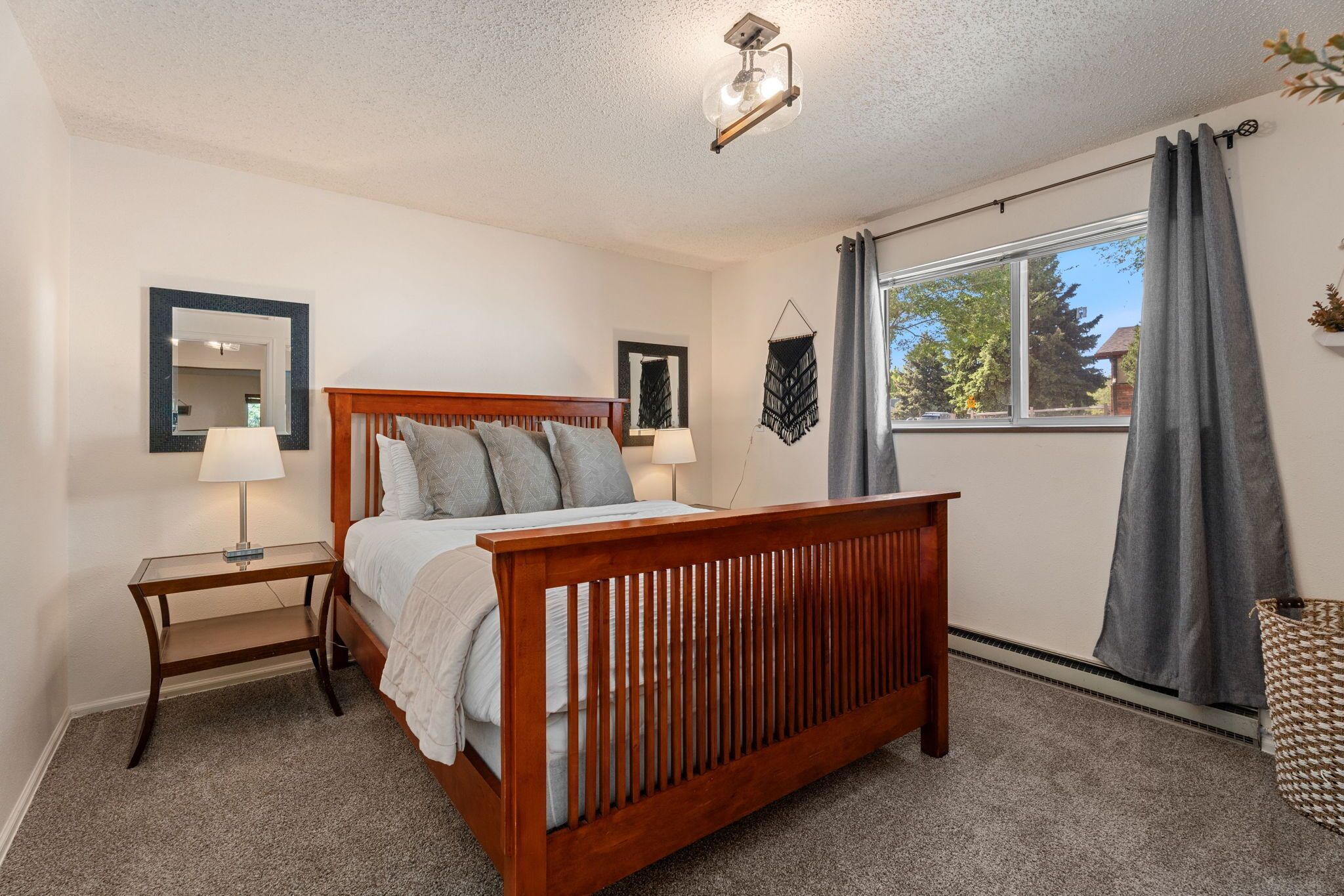 A bedroom with a wooden bed, two bedside tables with lamps, a large window with curtains, and a ceiling light fixture.