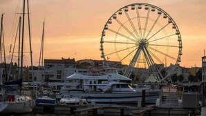 A marina with multiple boats docked, a large Ferris wheel, and buildings in the background. - Logement Entier : Cabane - Agde, France (Agde)