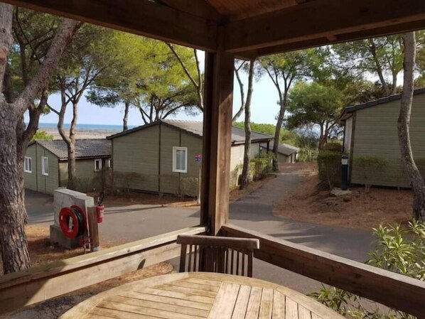 Un espace extérieur abrité, doté d’une table et de chaises en bois, entouré d’arbres et offrant une vue sur les maisons de plage.