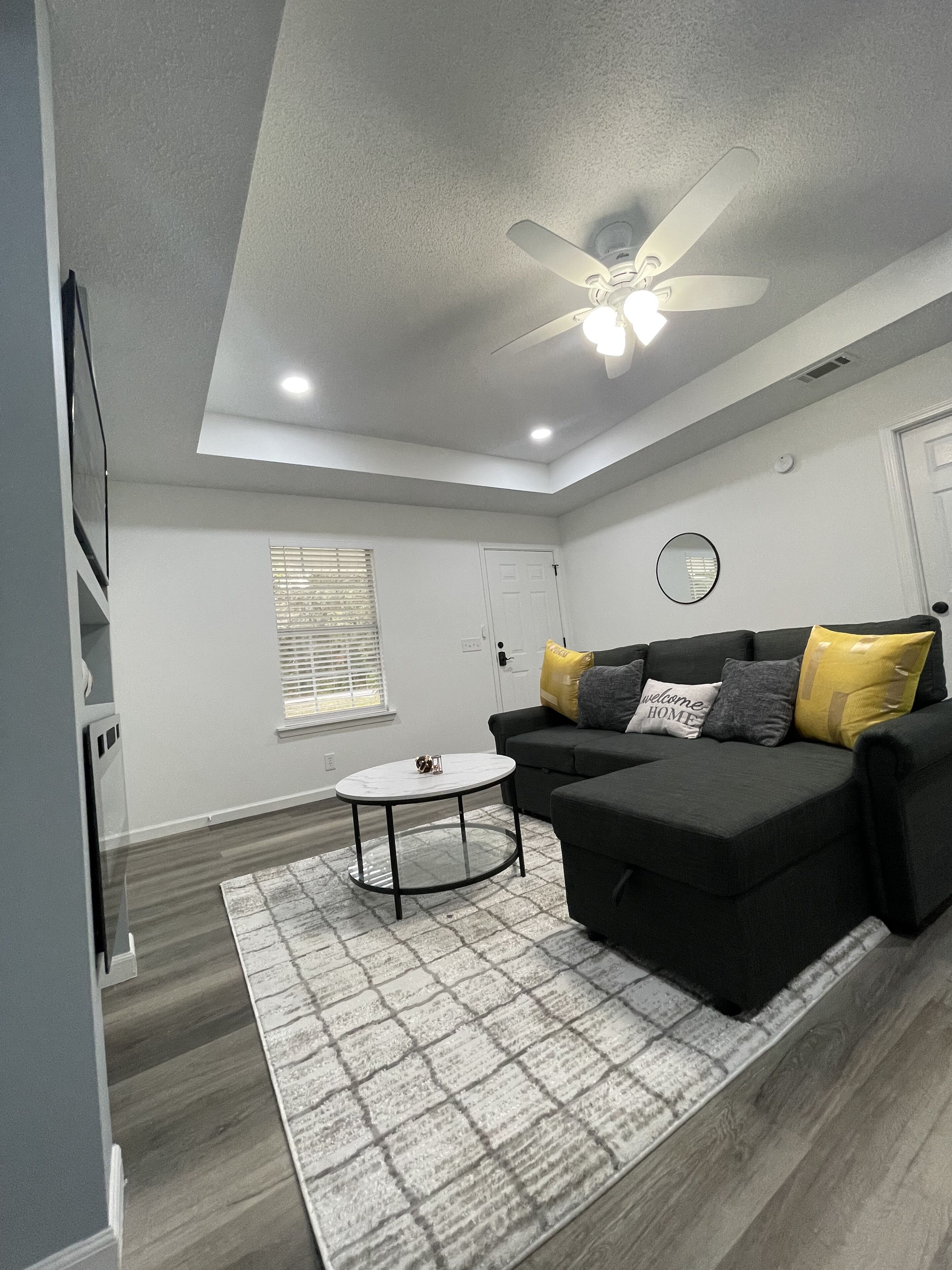 A modern living room with a ceiling fan, a black sofa with cushions, a small round table, and a patterned rug.