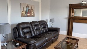 A living room with a dark brown leather sofa, a glass coffee table, a side table with a lamp, and a painting on the wall. - Newly renovated 2 bedroom apartment  (Saint John)