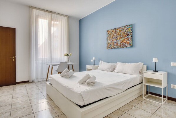 A hotel room with a bed, two bedside tables, a chair, a desk, and a painting on the wall.