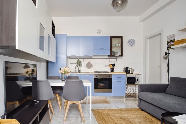 A modern kitchen with blue cabinets, a dining area with a white table and chairs, and a grey sofa in a living room.