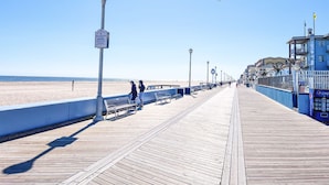 A wooden boardwalk with benches and street lamps, leading to a beach. - Oceanfront Boardwalk Condo Patio (Ocean City)