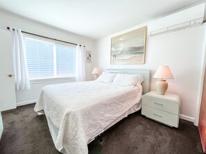 Une chambre à coucher avec un lit, une table de chevet, une lampe, une fenêtre avec des rideaux et un tableau au mur.