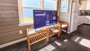 A living room with a flat-screen TV on a stand, a wooden chair, a kitchen area with a refrigerator, and large windows with blinds. - Oceanfront Boardwalk Condo Patio (Ocean City)