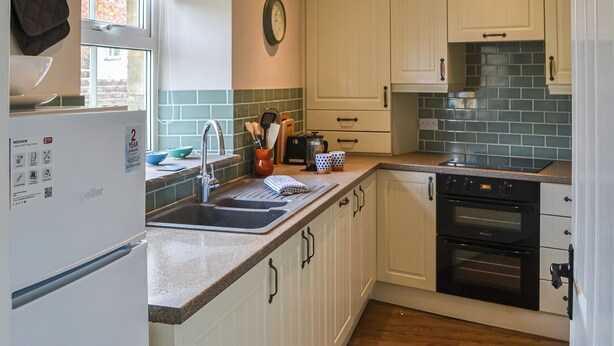 A kitchen with white cabinets, a stainless steel sink, a black oven, and a window with a view of trees. - 1 bedroom accommodation in Lesbury Near Alnwick (Lesbury Near Alnwick)