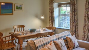 A cozy living room with a wooden dining table, two chairs, a sofa with patterned cushions, a floor lamp, and a window with curtains. - 1 bedroom accommodation in Lesbury Near Alnwick (Lesbury Near Alnwick)