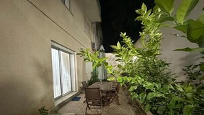 Een terras buiten met een tafel en stoelen, omgeven door planten en een gevel.