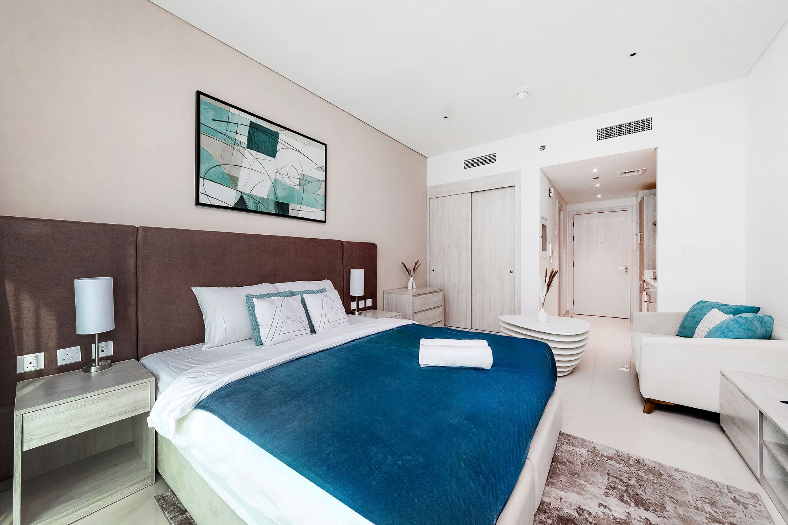 Una habitación de hotel moderna con una cama grande, una mesita de noche, un sofá blanco y una puerta que da a otra habitación.