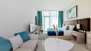 A modern hotel room with a sofa, a bed with a blue bedspread, a bedside table with a lamp, and a large window with curtains. - Modern studio on Palm Jumeirah (Dubai)