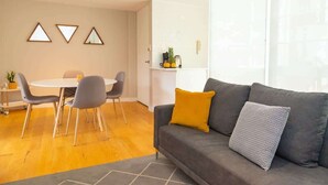 Ein modernes Wohnzimmer mit einem grauen Sofa, einem runden Esstisch mit Stühlen und einer Küchenzeile mit weißer Arbeitsplatte.
