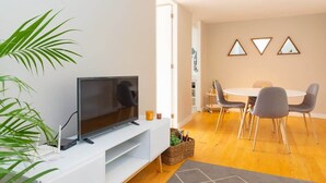 Ein modernes Wohnzimmer mit einem Flachbildfernseher auf einer weißen Konsole, einem Essbereich mit rundem Tisch und Stühlen sowie einer großen Topfpflanze.