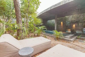 An outdoor seating area with a concrete structure overhead, surrounded by lush greenery and a small pool.