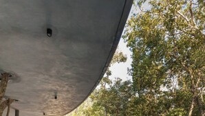 An outdoor swimming pool with lounge chairs under a concrete overhang, surrounded by trees.