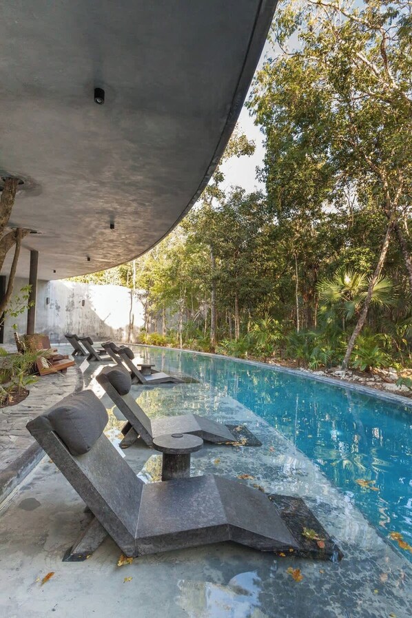 An outdoor swimming pool with lounge chairs under a concrete overhang, surrounded by trees.