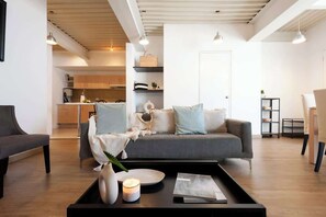 Een moderne woonkamer met een grijze bank, een salontafel en een eettafel. Achtergrond is een keuken met houten kastjes en een aanrecht.
