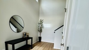 Un couloir avec un plancher en bois, un banc noir, un miroir, une plante en pot et une porte blanche.