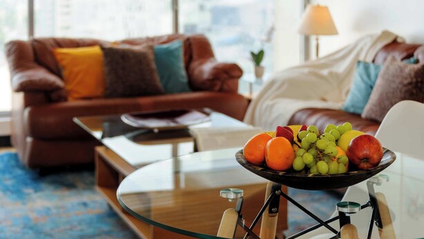 Un salon avec une table basse en verre, un canapé en cuir marron et un panier de fruits posé sur un présentoir.