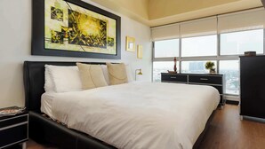 Une chambre à coucher moderne, dotée d’un grand lit, offrant une vue sur la ville grâce à de larges fenêtres, et ornée d’un tableau abstrait encadré au mur.