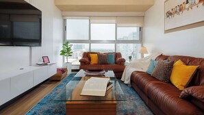 Un salon moderne avec un canapé en cuir marron, une table basse en verre, une console blanche et une grande fenêtre donnant sur la ville.