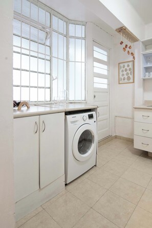 Une buanderie équipée d’un lave-linge, d’un évier et d’une fenêtre avec des stores.