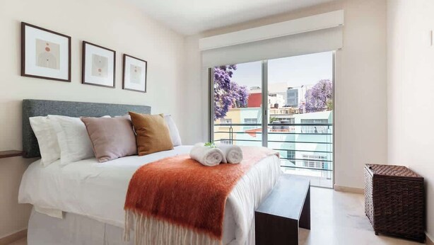 A bedroom with a bed, pillows, a blanket, a bench, a window, and a balcony.