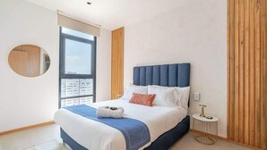 A modern bedroom with a large bed, a round mirror, a small side table, and a window with a city view.
