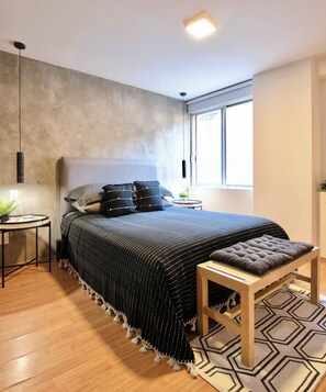 A modern bedroom with a large bed, a small bedside table, a potted plant, and a window with daylight.