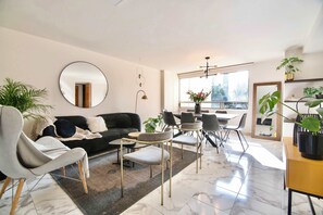 Una sala de estar moderna con un sofá negro, sillones grises, un espejo redondo y una zona de comedor con mesa y sillas.
