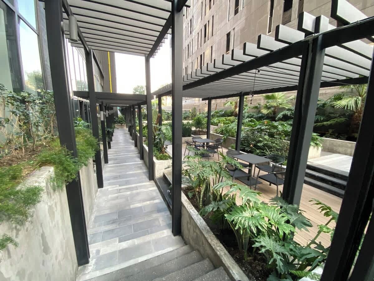 Un couloir extérieur abrité, avec des marches menant à un espace aménagé pour s’asseoir, entouré de verdure et avec un bâtiment en arrière-plan.