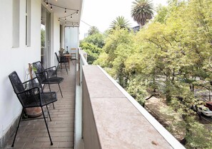 Un balcón con mobiliario exterior, vista a una calle arbolada y un edificio al fondo.