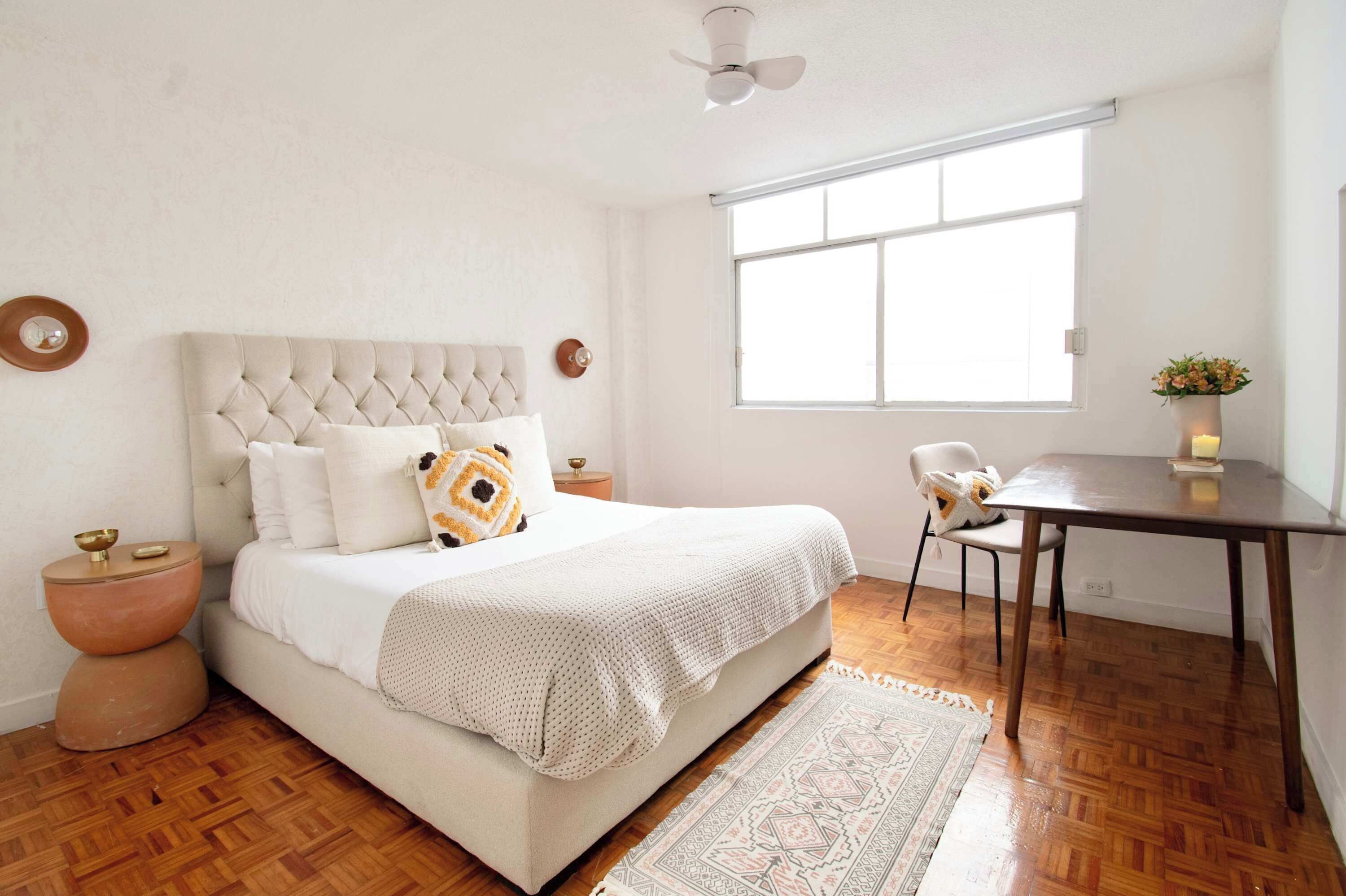 A bedroom with a bed, bedside tables, a chair, a table, a window, and a ceiling fan.
