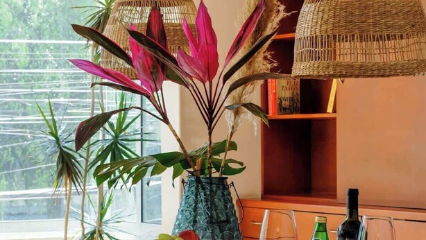Een eethoek met een houten tafel, rieten stoelen, een vaas met planten en wijnglazen.
