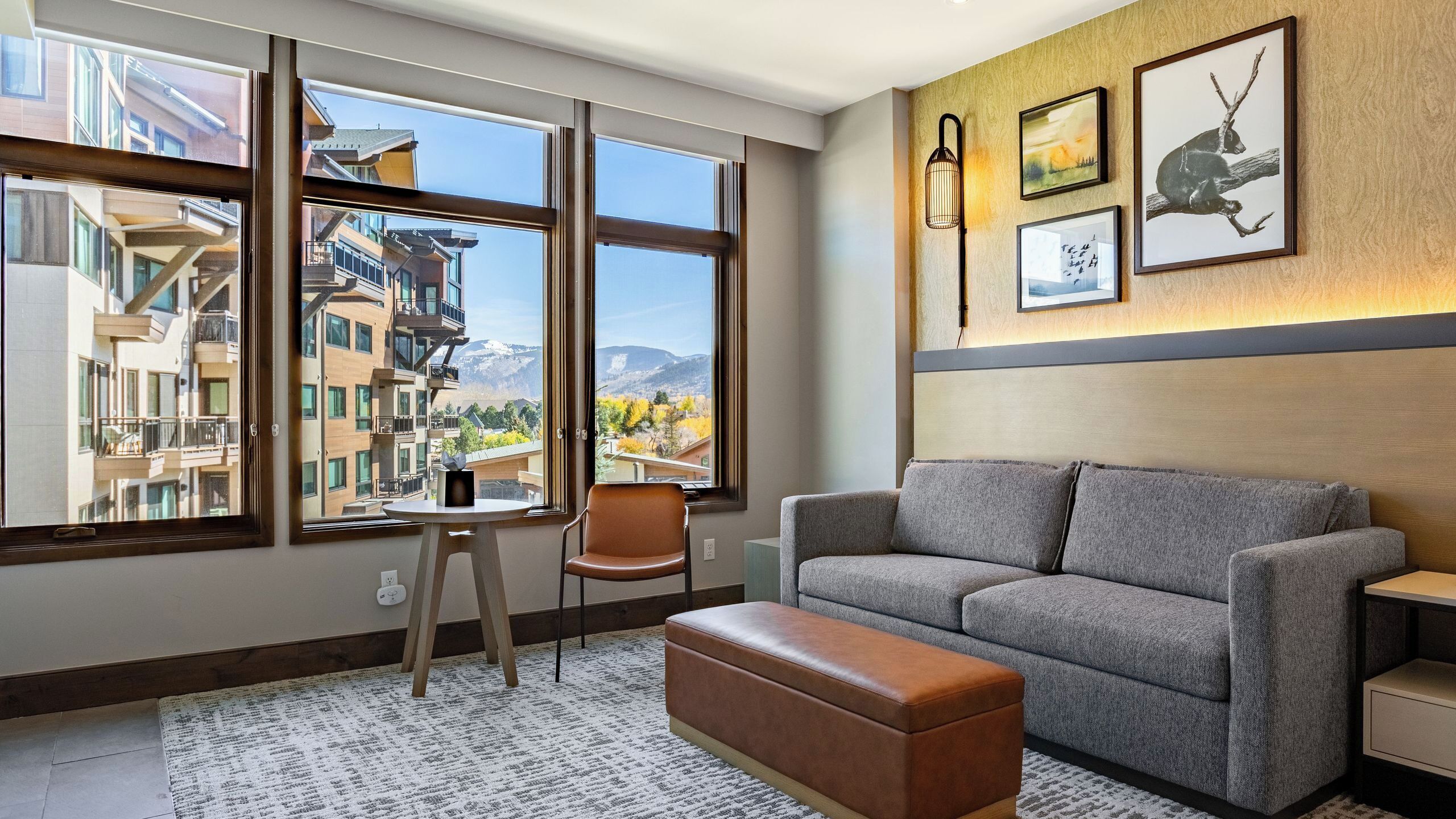 Ein modernes Wohnzimmer mit einem grauen Sofa, einem braunen Hocker, einem Holzstuhl und einem Tisch. Große Fenster bieten Blick auf ein Gebäude und Berge.