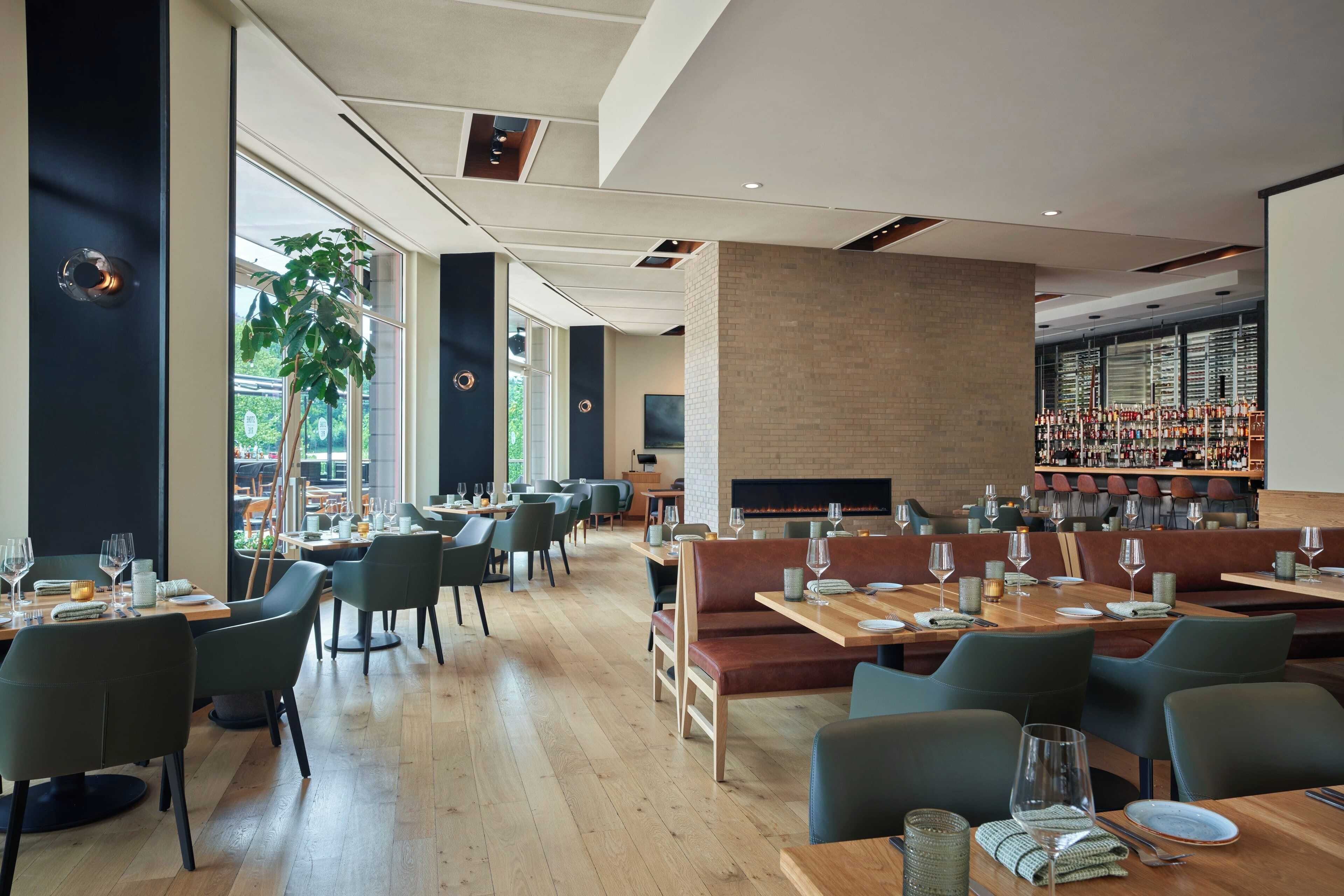 A modern restaurant with wooden tables and green chairs, a brick wall, and a bar area with shelves of bottles.
