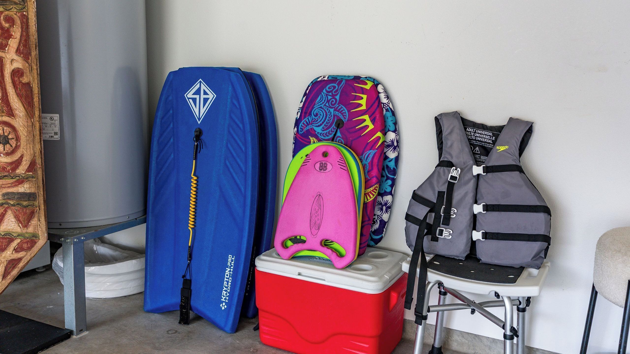 A room with a red cooler, a blue bag, a pink bag, and a gray bag on a metal stand.