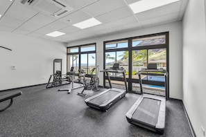 A gym with treadmills, exercise bikes, and weight machines, large windows with a view of a pool and palm trees. - Pakele Oahu by Avantstay 5 Min to Makaha Beach (Waianae)