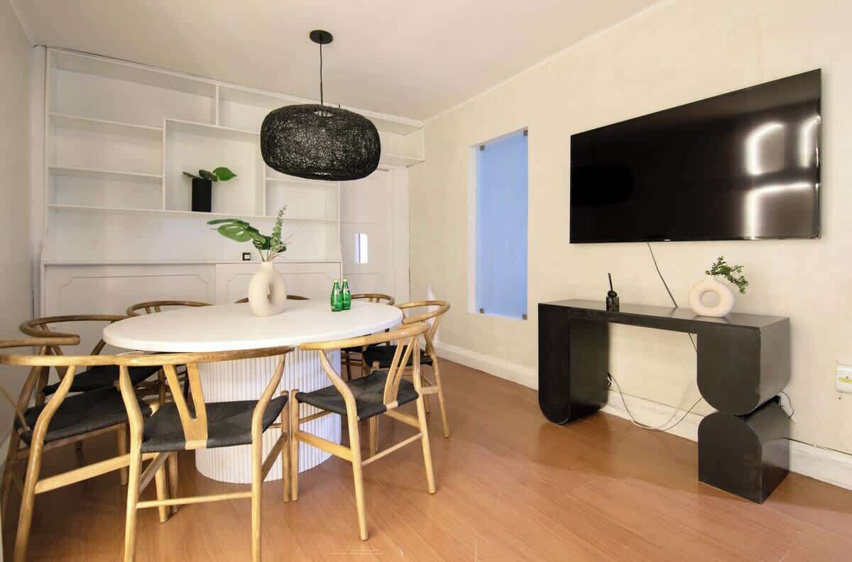 A modern dining area with a round table, wooden chairs, a black pendant light, and a TV mounted on the wall.