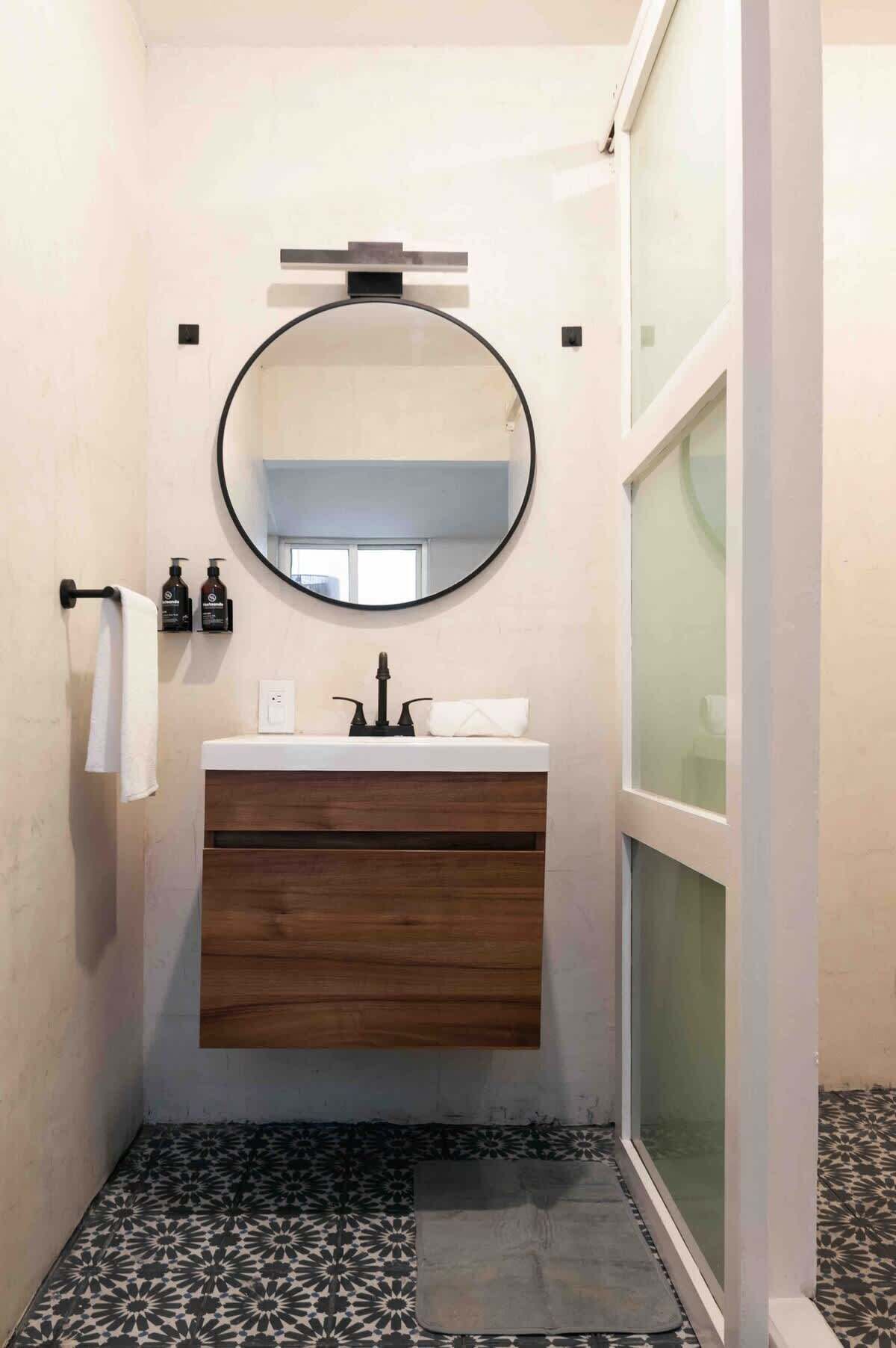 A bathroom with a round mirror, wooden vanity, two bottles of shampoo, a towel, and patterned floor tiles.
