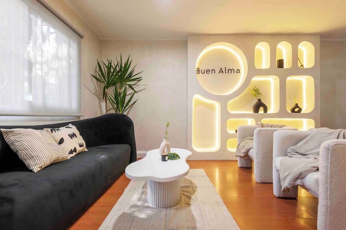 A modern living room with a black sofa, a white coffee table, and a wall with illuminated geometric shapes and the text "Buen Alma".