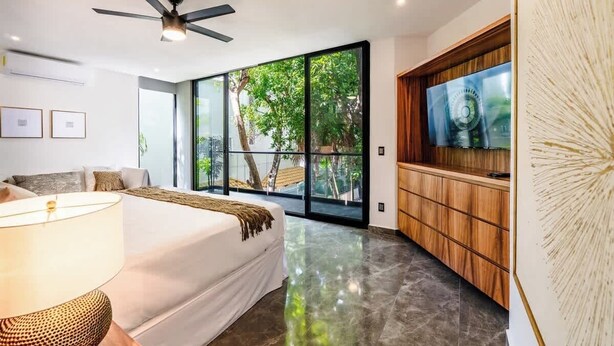 A modern bedroom with a large bed, a ceiling fan, a TV on a wooden cabinet, and a view of greenery through large windows. - Stunning modern villa with pool & beach access (Playa del Carmen)