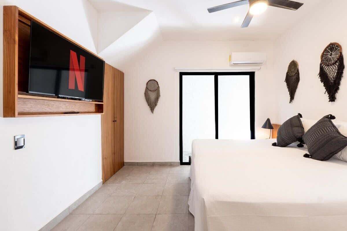 A bedroom with a bed, a TV mounted on the wall, a ceiling fan, and decorative wall hangings.