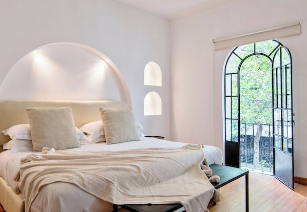 A bedroom with a large bed, a small table, and a view of greenery through a window.
