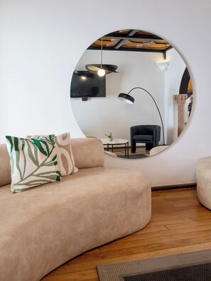 A modern living room with a round mirror, a beige sofa, a black armchair, a floor lamp, and a television mounted on the wall.
