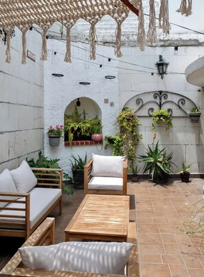 An outdoor patio with a wooden bench, a table, and a sofa with cushions. There are potted plants and hanging plants on the walls.