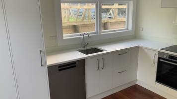 A modern kitchen with white cabinetry, a built-in dishwasher, a double sink, and a window overlooking a wooden deck.
