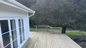 Una terraza de madera con barandilla, un edificio blanco con puertas de vidrio y un árbol al fondo.